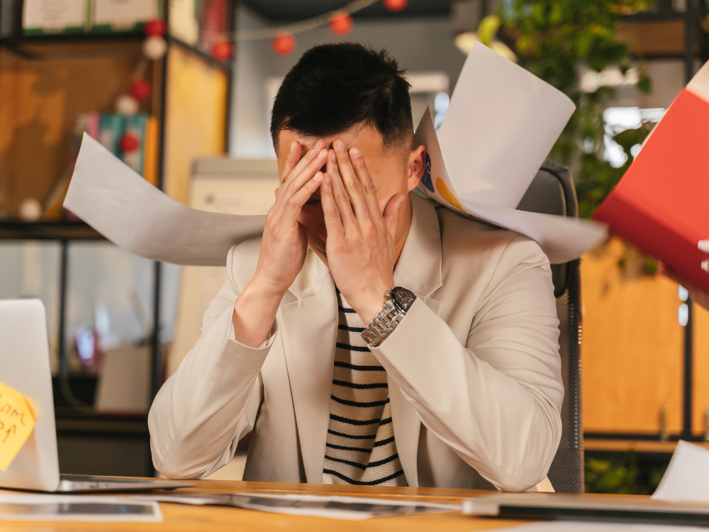 Stress beheersen met Hypnotherapie 9 Man aan een bureau met zijn handen voor zijn gezicht, omringd door papieren.